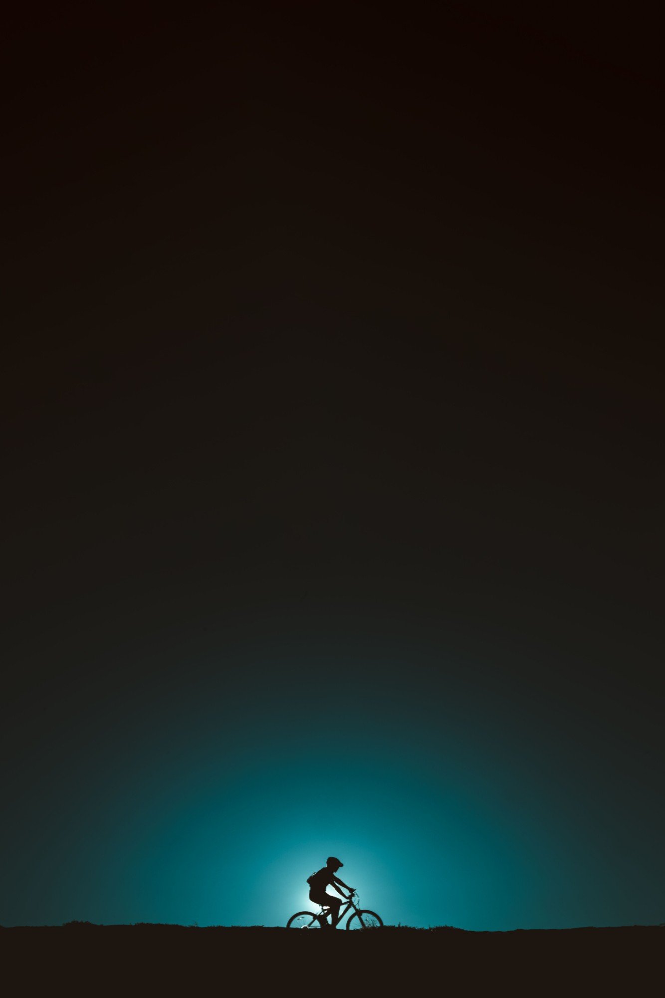 Silhouette of a cyclist on a ridge against a teal glow in the night sky