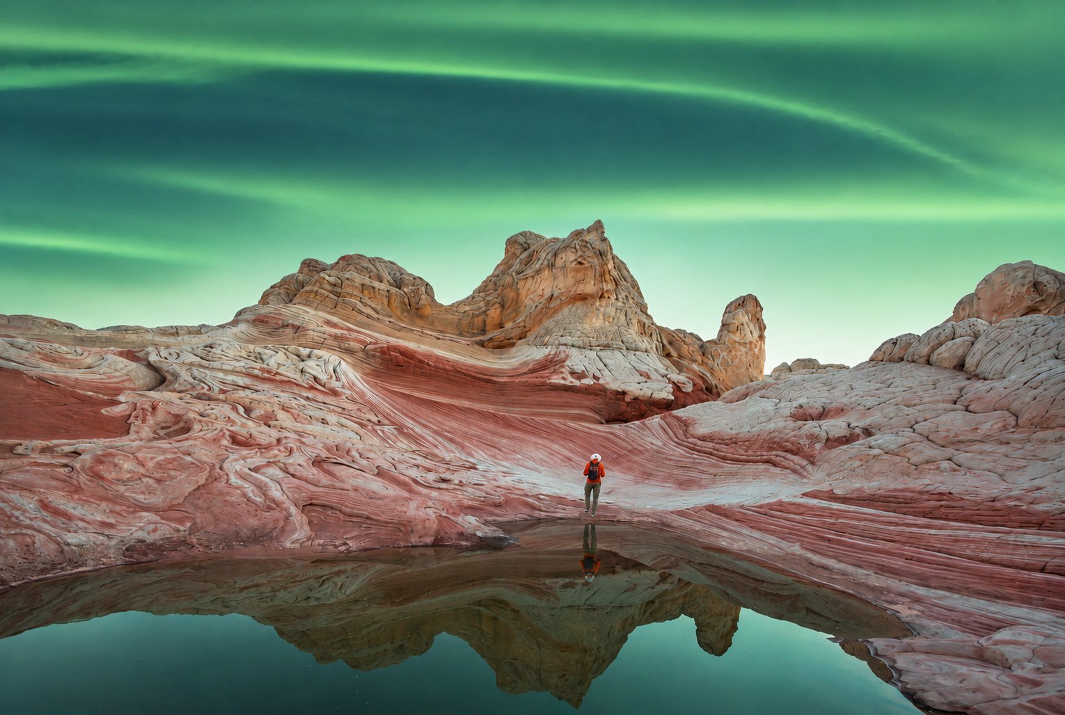 A lone walker on wave-shaped sandstone under a green aurora, reflected in still water
