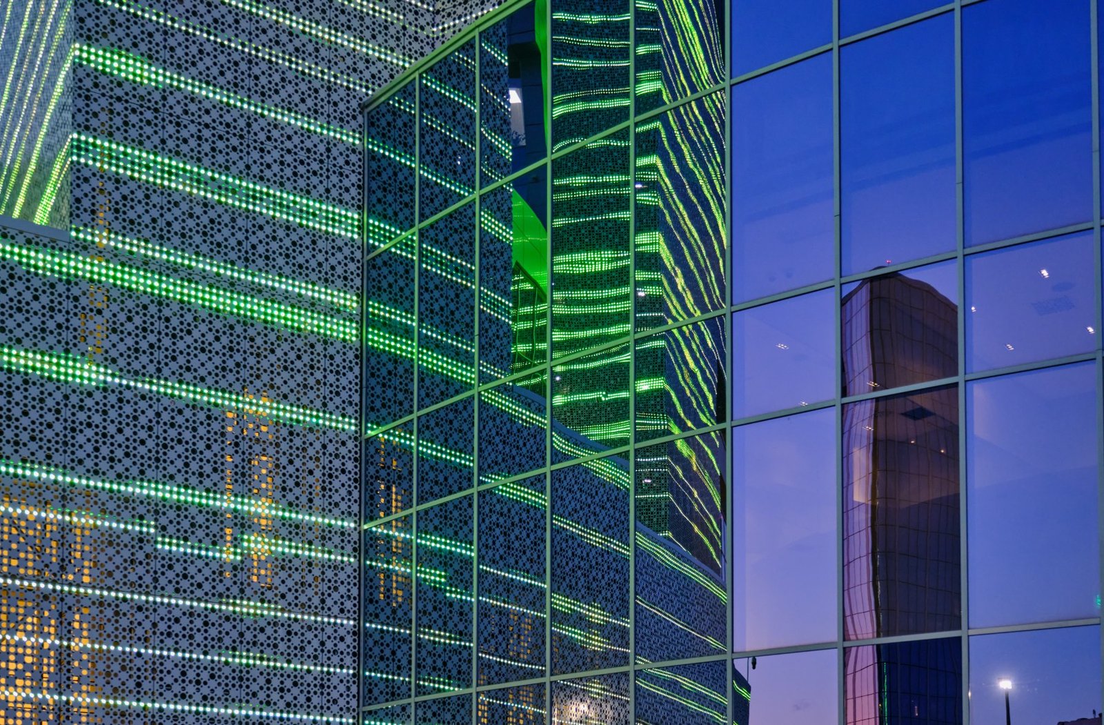 Glass facade lit with green LED lines at dusk, reflecting a neighboring tower